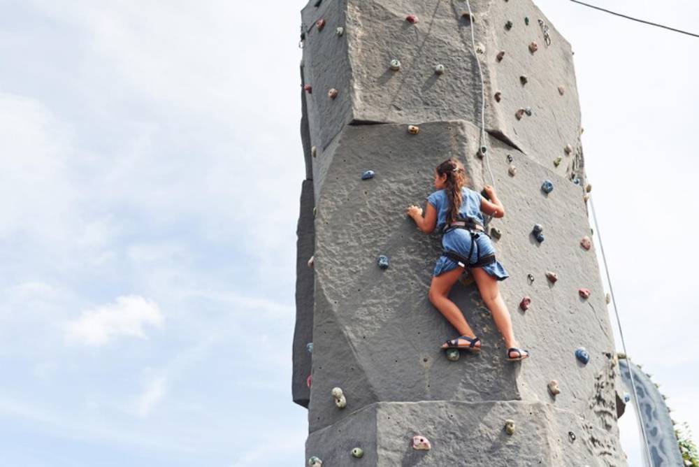A young girl climbs a rock wall, wearing a harness and blue attire, with a clear sky in the background.