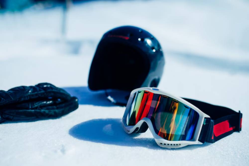 A black ski helmet, colorful ski goggles, and black gloves resting on fresh snow, ready for a day on the slopes.