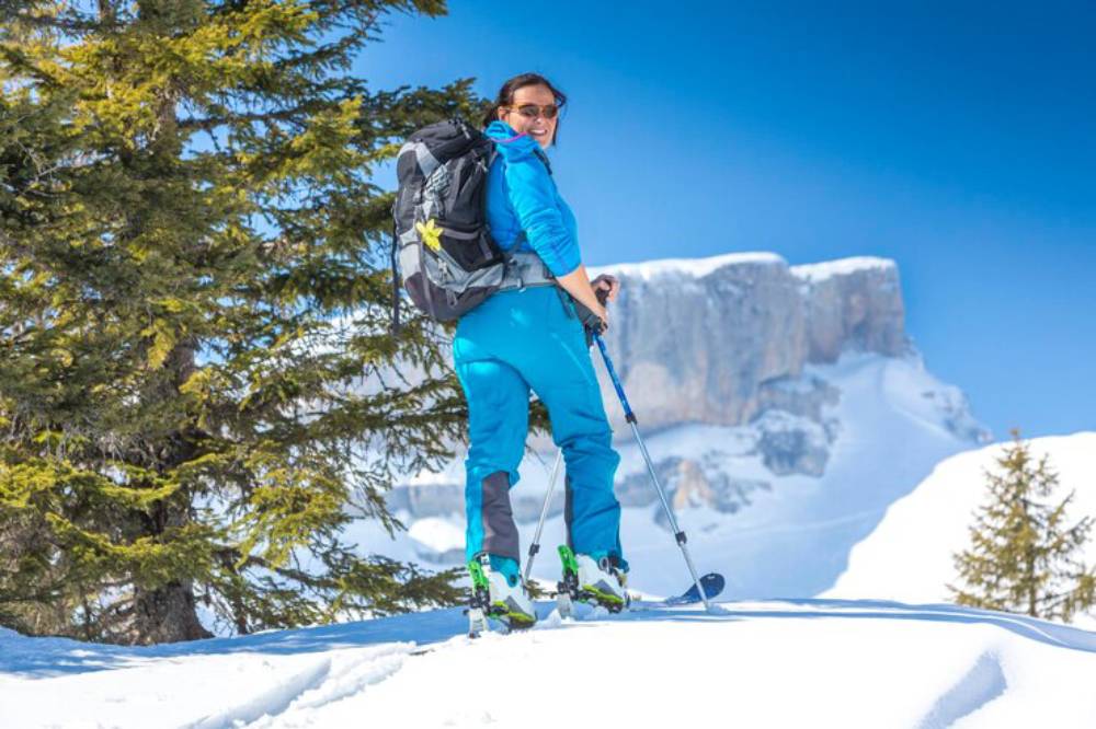 A skier in a bright blue outfit stands on a snowy mountain trail, surrounded by tall trees and majestic peaks under a clear blue sky.