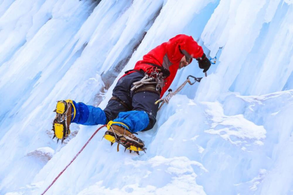 A climber in a red jacket ascends an icy wall using ice tools and crampons, surrounded by blue icicles and snow.