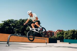A BMX rider performs a trick on a rail at a skatepark, surrounded by greenery and colorful buildings under a clear blue sky.