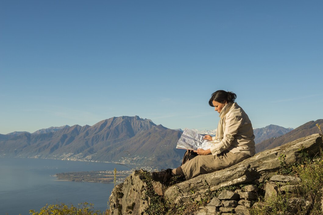 A hiker sits on a rocky ledge, studying a map, surrounded by mountains and a lake under a clear blue sky.