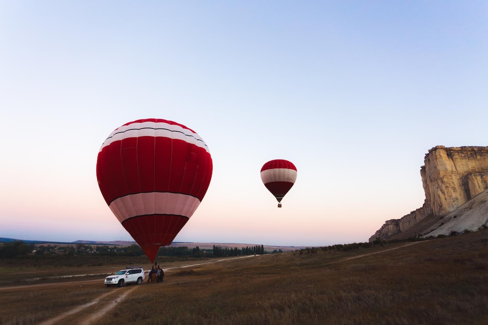 A white SUV parked on a dirt path with two red and white hot air balloons soaring above a scenic landscape at dusk.