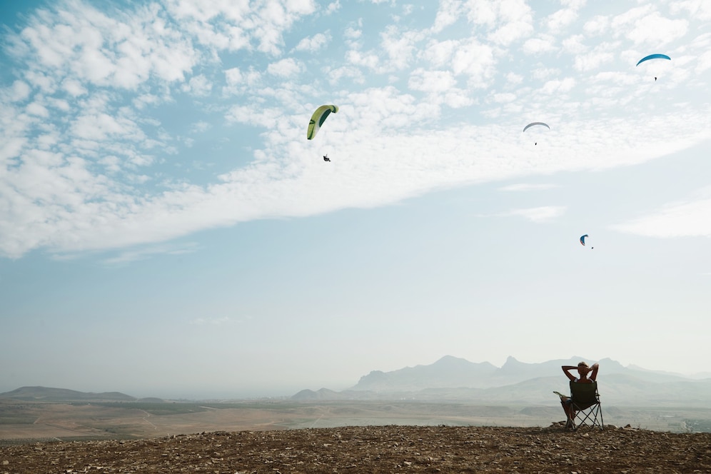A person relaxes in a chair, watching paragliders soar above a vast landscape with mountains under a partly cloudy sky.