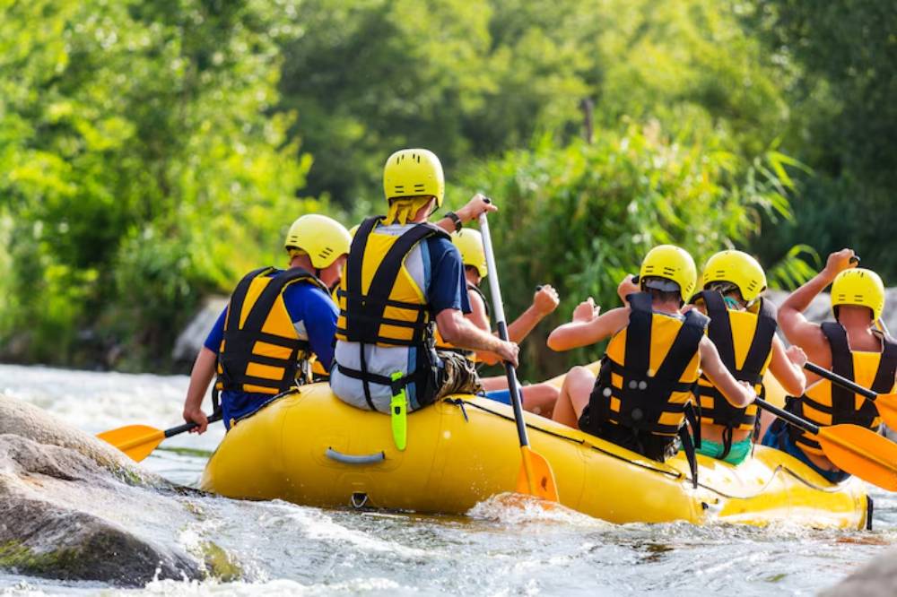 A group of six people in yellow helmets and life jackets paddling a yellow raft through a river surrounded by lush greenery.