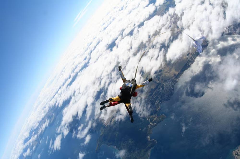 A tandem skydiver descends through fluffy clouds, with a view of land and water below, capturing the thrill of freefalling from above.
