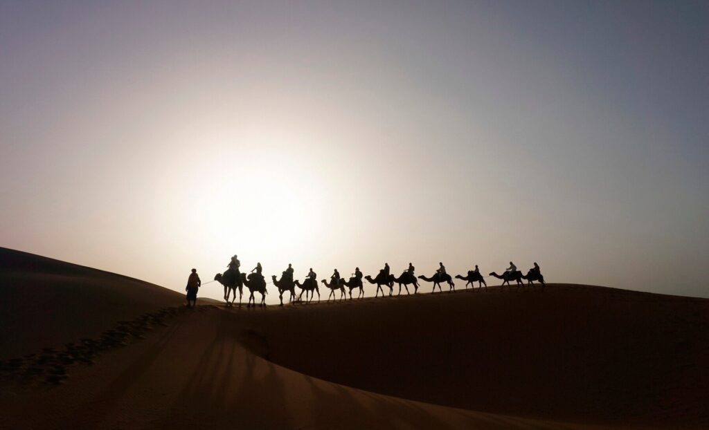 Silhouetted figures on camels trek across a sand dune against a glowing sunset sky in a serene desert landscape.