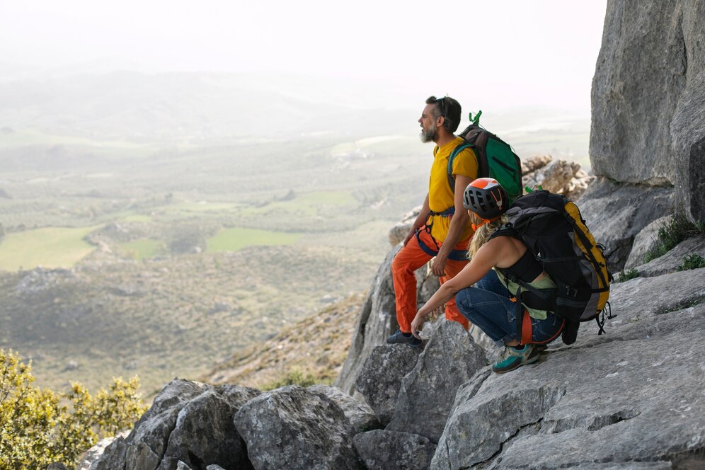 Two climbers resting on a rocky ledge, overlooking a lush green valley, equipped with backpacks and climbing gear.