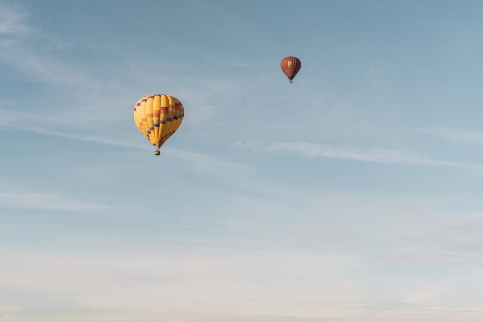 Two colorful hot air balloons float high in a clear blue sky, surrounded by soft wisps of white clouds.