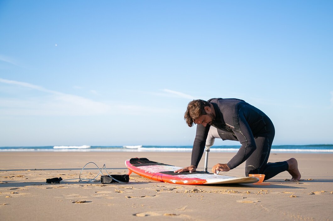A person kneels on the sand, preparing their surfboard, with waves gently lapping in the background under a clear blue sky.