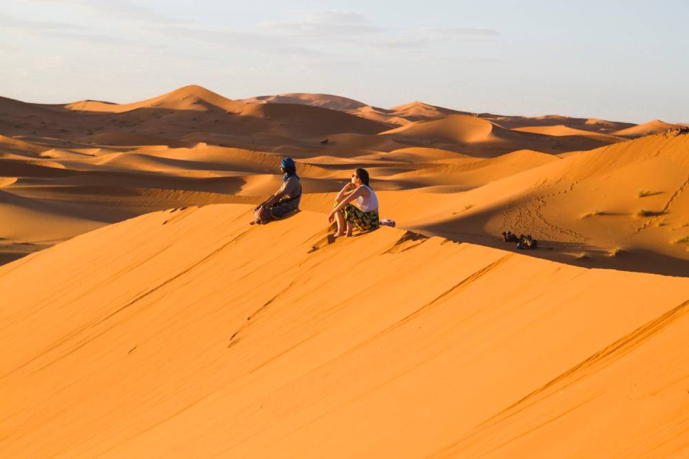 Two people sit atop a golden sand dune in a vast desert, enjoying the serene landscape and soft evening light.