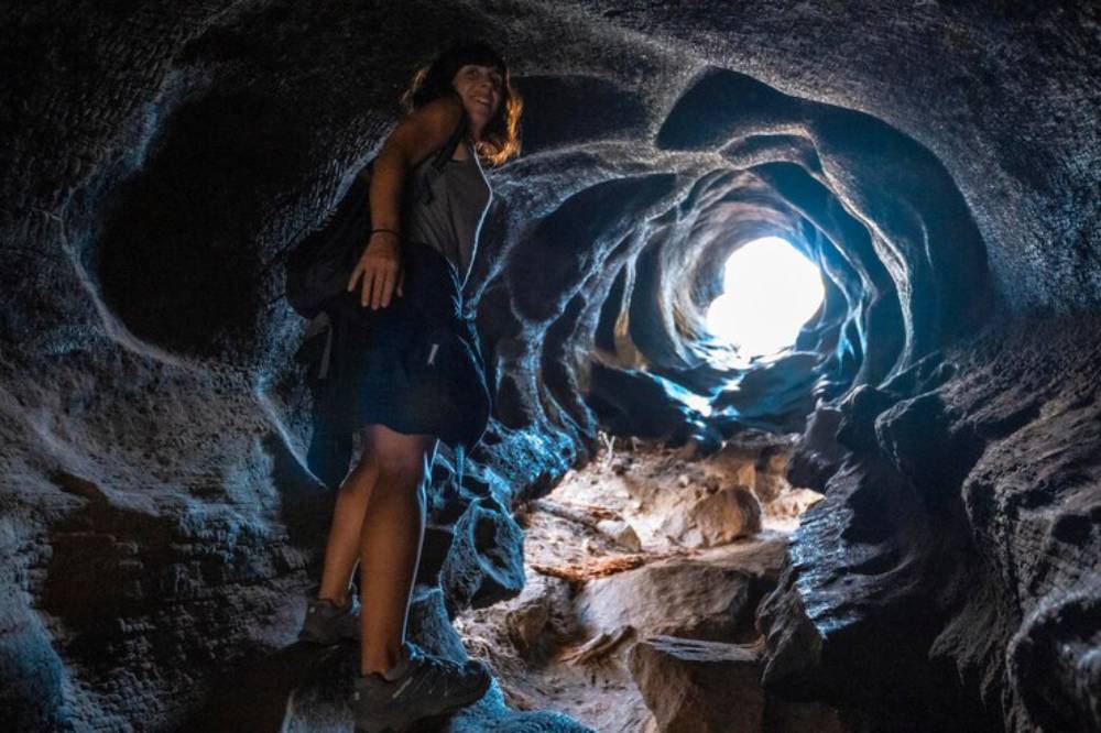 A person stands inside a dark cave, illuminated by bright light at the entrance, showcasing textured rock formations and shadows.