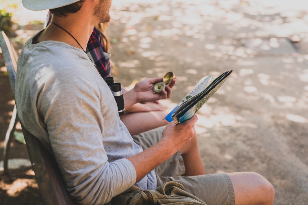 A man with a binocular around his neck holds a compass and a map while sitting on a bench beside a woman in a plaid shirt.