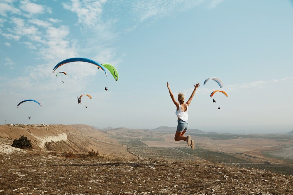 A woman jumps joyfully on a hilltop, surrounded by colorful paragliders soaring against a clear blue sky.