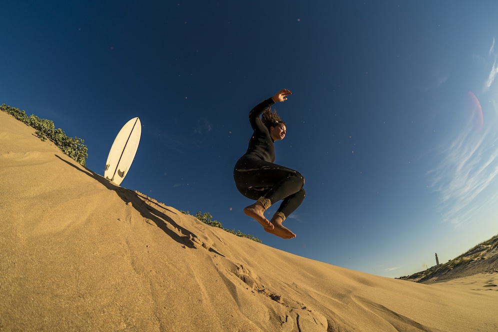 A person in a wetsuit leaps off a sand dune with a surfboard resting nearby, under a clear blue sky.