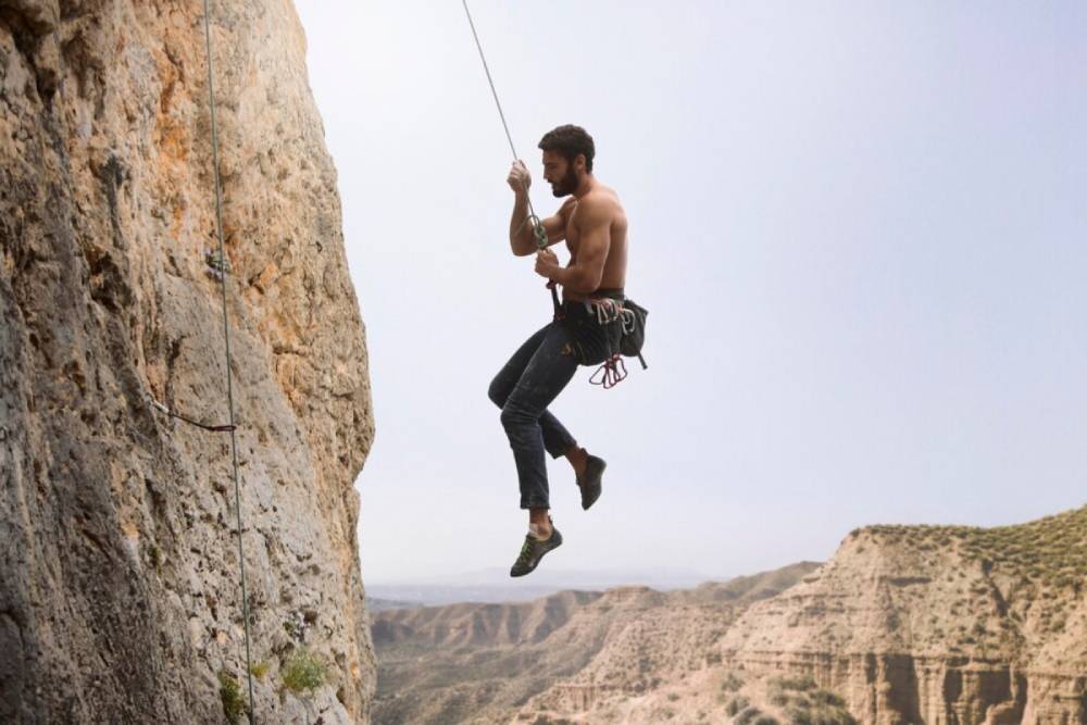 A shirtless climber hangs mid-air from a rock face, gripping a rope, with rugged cliffs and a vast landscape in the background.