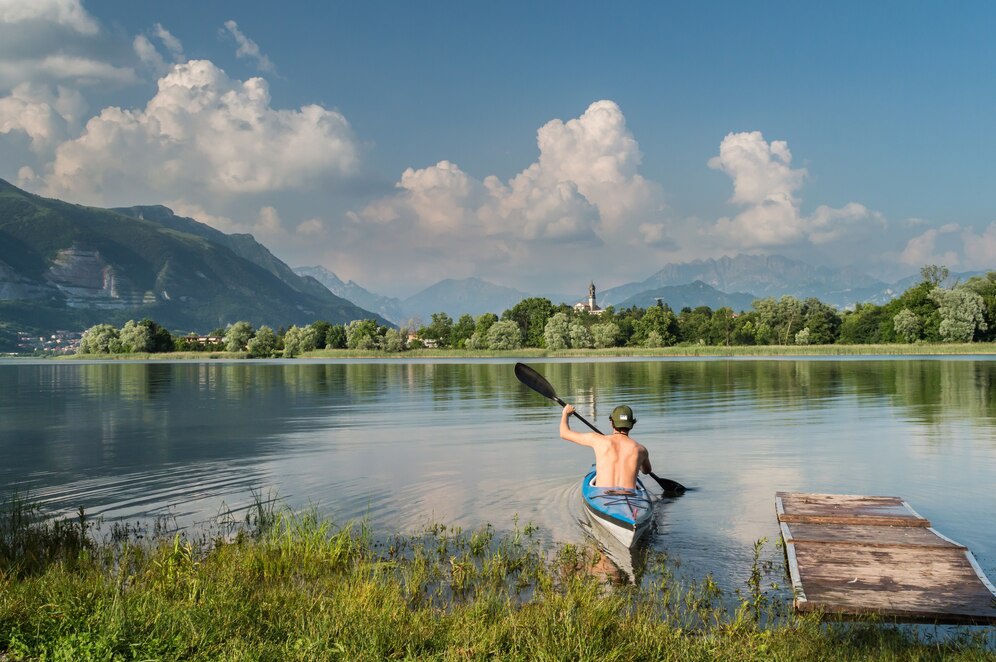 A shirtless man paddles a kayak on a calm lake, surrounded by lush greenery and mountains under a partly cloudy sky.