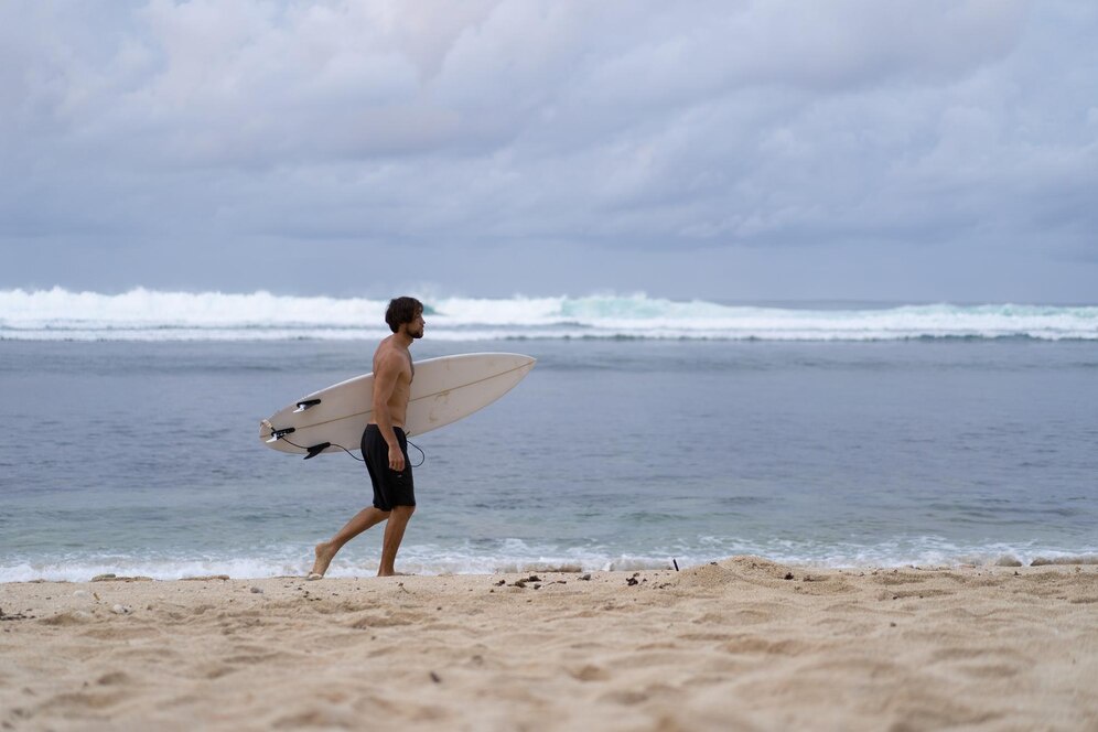 A shirtless surfer walks along the sandy beach, holding a surfboard, with waves rolling in under a cloudy sky.