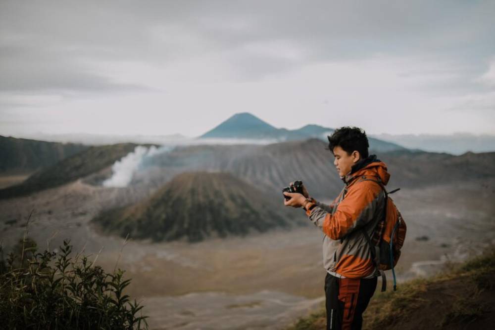 A person stands on a volcanic landscape, holding a camera, with a distant smoking crater and mountains under a cloudy sky.