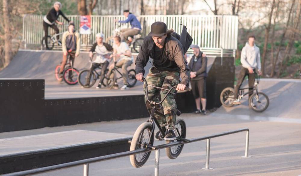 A BMX rider performing a trick on a rail at a skatepark, surrounded by other riders and spectators in a sunny outdoor setting.