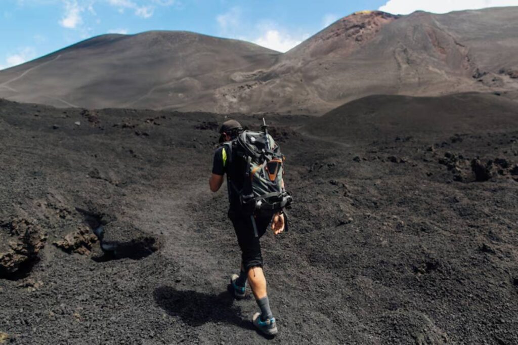 A hiker traverses a rugged volcanic landscape, with dark, rocky terrain and mountains under a blue sky.