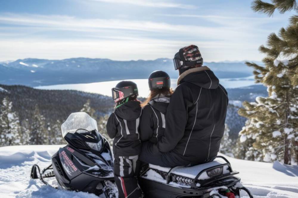 A family of three, wearing winter gear, enjoys a scenic view of snow-covered mountains and a lake while sitting on a snowmobile.