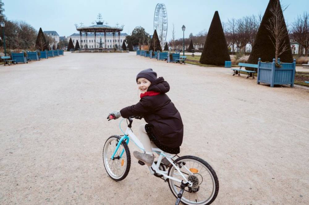 A child in a winter coat rides a white bicycle in a spacious park with neatly trimmed hedges and a large Ferris wheel in the background.