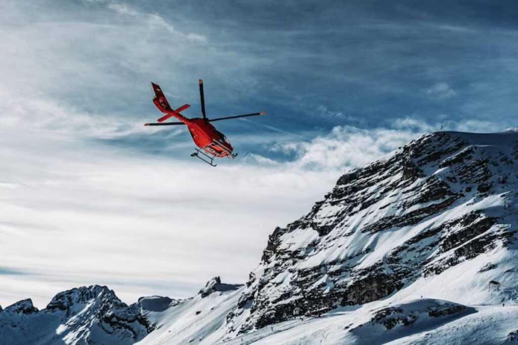 A red helicopter hovers above snow-covered mountains under a dramatic sky.