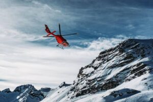 A red helicopter hovers above snow-covered mountains under a dramatic sky.