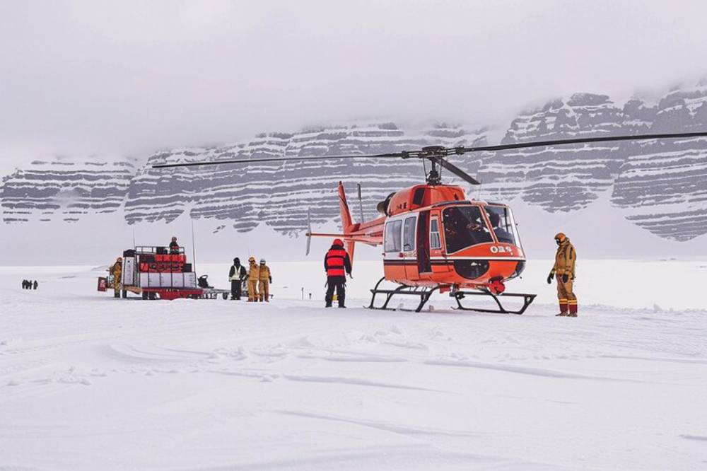 Persons near a helicopter on a snowy landscape, with steep mountains in the background, during a cold and cloudy day.