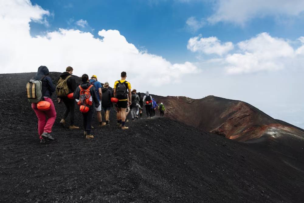 A group of hikers, wearing helmets and backpacks, ascend a volcanic landscape under a cloudy blue sky.
