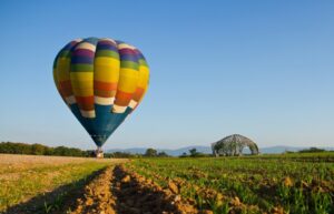 A colorful hot air balloon prepares for flight over a green field, with a unique arch structure visible in the distance under a clear blue sky.
