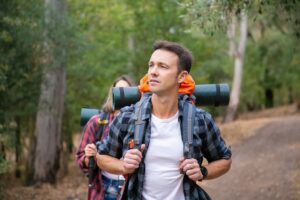 Two hikers with backpacks and sleeping mats walk along a forest trail, surrounded by lush greenery and trees.