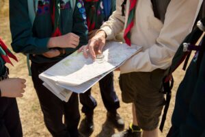 A group of young scouts gathers around an adult leader who points at a map while holding a compass, engaging in navigation training.