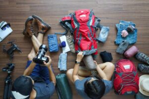 Two people organizing travel gear on a wooden floor, including clothing, a red backpack, and photography equipment.