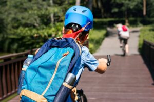 A boy in a blue helmet rides his bike along a wooden path, wearing a backpack and holding the handlebars, with another cyclist in the distance.