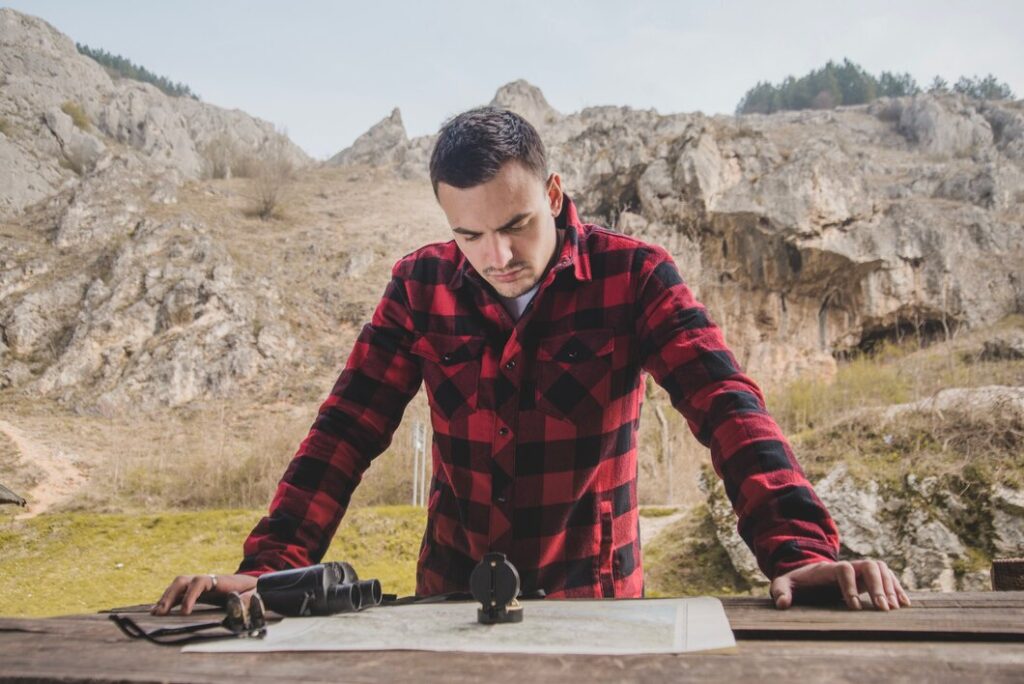 A person in a red and black checkered shirt studies a map on a wooden table, surrounded by rocky terrain and greenery in the background.