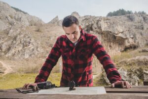 A person in a red and black checkered shirt studies a map on a wooden table, surrounded by rocky terrain and greenery in the background.