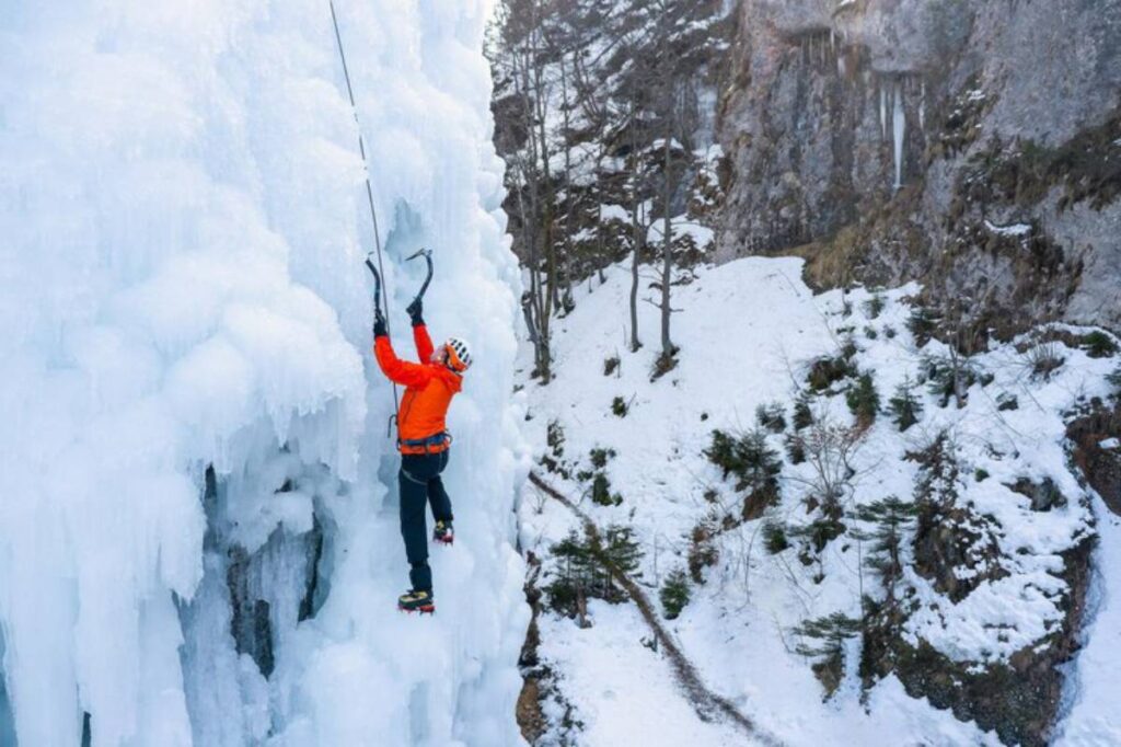 A climber in an orange jacket ascends a frozen icefall, using ice axes and crampons, surrounded by snow and trees.