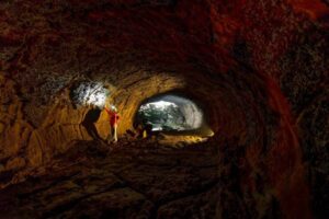 A person in a red jacket stands inside a cave, illuminated by flashlight, showcasing the cave's textured walls and distant opening.