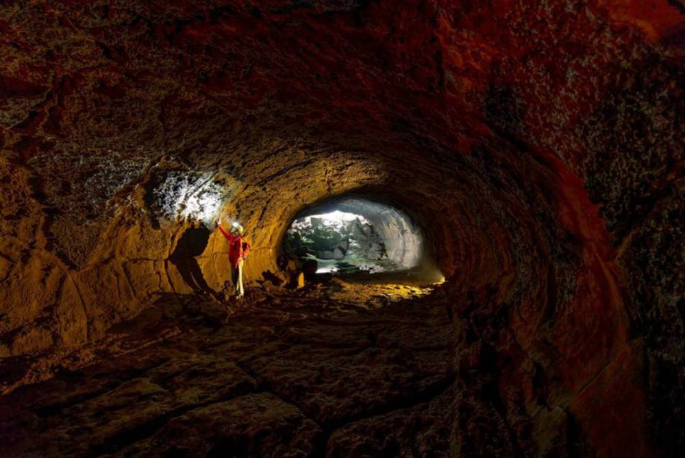 A person in a red jacket stands inside a cave, illuminated by flashlight, showcasing the cave's textured walls and distant opening.
