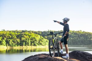 A cyclist in a helmet gestures towards a scenic lake surrounded by lush greenery, standing beside a mountain bike on rocky terrain.