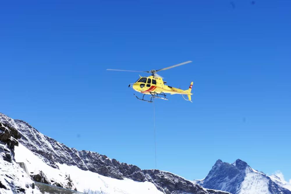 A yellow helicopter hovers above snow-capped mountains under a clear blue sky, with a cable hanging from its fuselage.