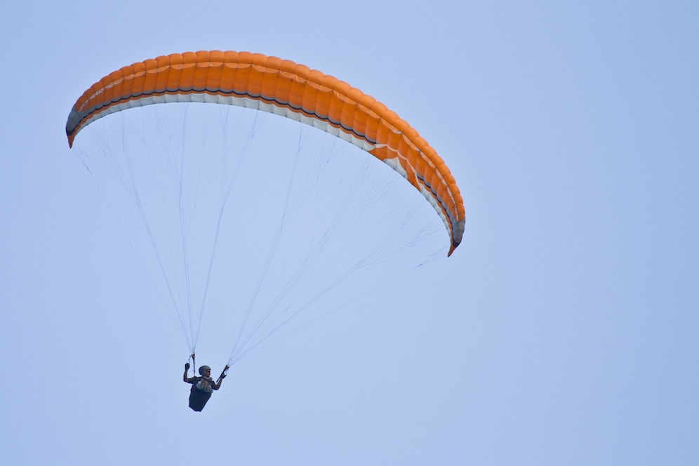 A person paraglides against a clear blue sky, soaring beneath a vibrant orange and white parachute.