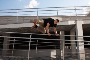 A person performs a fluid parkour move, swinging over a railing on a modern building's upper level under a clear blue sky.