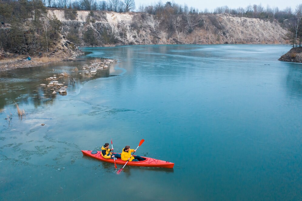 Two people in yellow jackets paddle a bright red kayak on a tranquil blue lake surrounded by rocky cliffs and sparse trees.