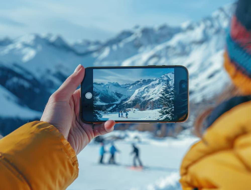A person in a yellow jacket holds a smartphone, displaying a snowy mountain scene with skiers, capturing the beautiful winter landscape.