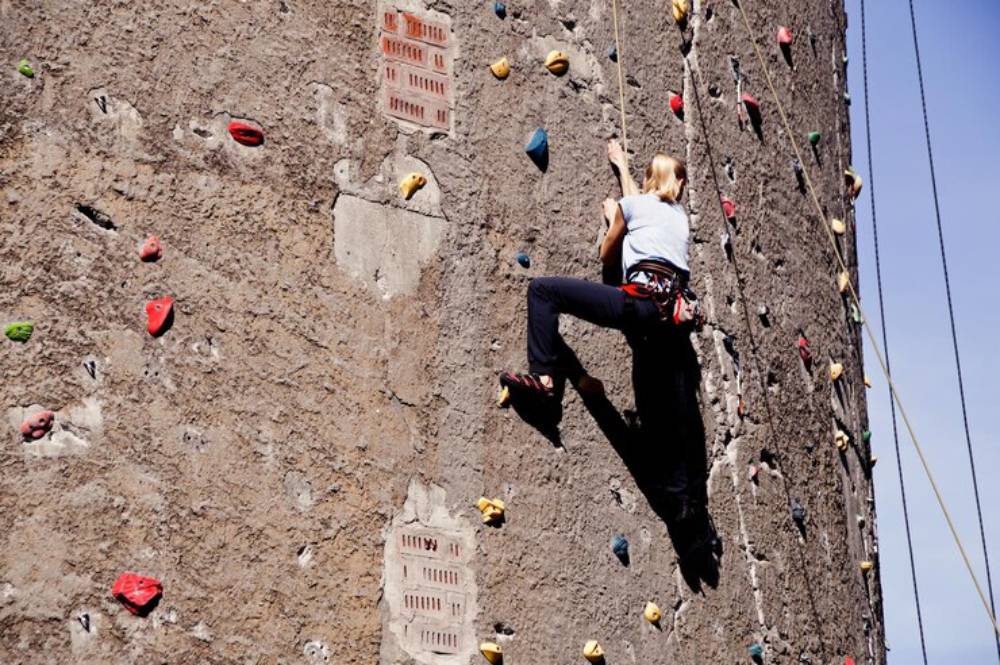 A person climbs a textured rock wall dotted with colorful holds, secured with a harness and rope against a clear sky.