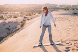 A person stands on a snowboard, poised on a sandy dune at sunset, surrounded by gentle hills and sparse vegetation.