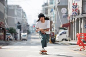 A skateboarder in a white shirt and green pants performs a trick on an empty street, surrounded by urban buildings and parked cars.